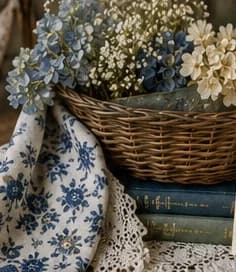 A woven basket of blue hydrangeas and baby's breath resting on old books beside blue floral fabric